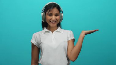 Kulaklık takan bir kadın, parlak yeşil bir arkaplanı işaret ederken gülümseyerek sunum ya da ürün yerleştirme önerisinde bulunuyor..