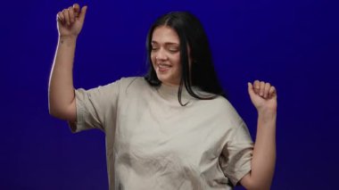 Uzun saçlı bir kadın neşeyle dans ediyor, parlak mavi bir arka planda, gösteriş yapıyor, moda ve ifade tarzını gösteriyor.