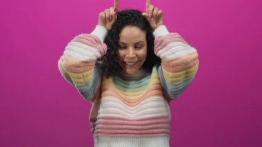 Renkli süveter giyen İspanyol kadın parlak pembe arka plana karşı şakacı bir şekilde boynuz hareketi yapıyor.