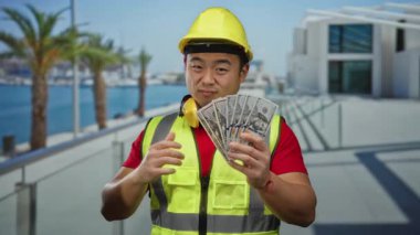 Deniz kenarındaki gezinti güvertesinde güvenlik yeleği ve başlık takan genç bir adam bir elinde baş parmak, diğer elinde dolarlarla bize yardım ediyor..