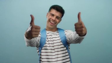 Mavi bir arkaplan önünde başparmağıyla gülümseyen genç adam, çizgili bir gömlek ve sırt çantası giyiyor, pozitifliğini ve güvenini ifade ediyor..