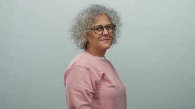 Kıvırcık gri saçlı, gözlüklü ve pembe gömlekli yaşlı bir kadın, izole edilmiş beyaz arka plan duvarında sıcak bir gülümsemeyle bilgelik ve rahatlık iletiyor..