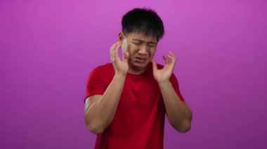 Kırmızı tişörtlü genç Çinli adam, iyi aydınlatılmış bir stüdyo ortamında, izole edilmiş pembe bir duvar arkasına karşı korkmuş duygularını ifade ediyor..