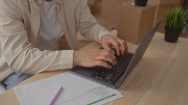 Yeni bir evde dizüstü bilgisayar üzerinde çalışan beyaz bir adam. Etrafı karton kutular, ev bitkileri ve ofis malzemeleriyle çevrili..