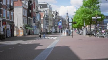 Parlak bir gökyüzü altında tarihi mimari ve ağaçlarla Avrupa 'nın bir caddesini geçen insanların bulanık görüntüsü.