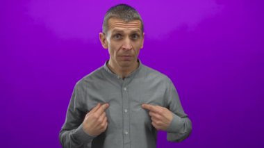 Kafkasyalı bir adam kendini parlak mor bir arka plana işaret ediyor, gri bir gömlek giyiyor, izole edilmiş ve jestinde anlamlı..