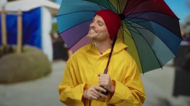 Sarı yağmurluklu yakışıklı bir adam, mavi gökyüzünün altında neşeli bir ifadeye sahip şehir caddesinde renkli bir şemsiye tutuyor, parlak bir günde kırmızı bir bere ve günlük kıyafetler giyiyor..