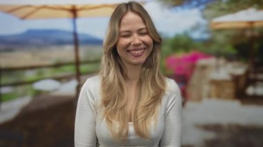 Sarışın kadın, güneşli bir restoran terasında gülümserken elleriyle yanaklarını tutar; neşe, neşe, dinginlik..