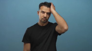 Siyah tişörtlü genç İspanyol adam mavi arka plana karşı duruyor düşünceli ifadeler ve jestler sergiliyor..