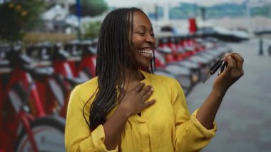 Parlak sarı tişörtlü gülümseyen bir kadın arka planı bulanık, kiralık bisikletlerin yanında duran araba anahtarlarını tutuyor..