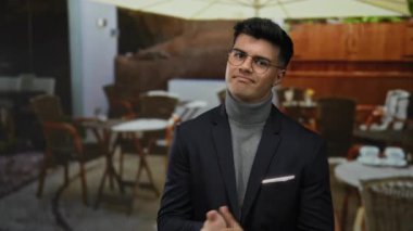 Şehir restoranındaki terasta duran gözlüklü genç adam, dışarıda masa ve şemsiyenin altındaki sandalyelerle yakışıklı ve kendinden emin görünüyor..