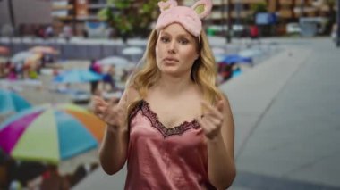 Gece elbiseli, tavşan kulaklı bir kadın renkli bir plaj arkaplanının yanında konuşuyor, gündelik ve genç yaz atmosferini vurguluyor..