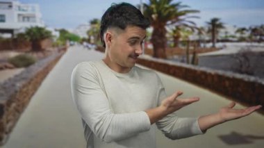 Genç ispanyol adam neşeli bir deniz kenarında yürüyüş yolunda palmiye ağaçlarıyla neşeli bir şekilde el kol hareketi yapar, açık deniz kenarında günlük açık süveter giyer.