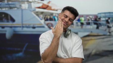 Sahilde bir teknenin yanında boynunda kulaklıklarla gülümseyen bir adam, arka planda insanlarla birlikte dışarıdaki bir ortamda samimiyet ve dostluk yayıyor..