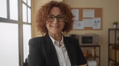 Siyah ceketli İspanyol kadın, parlak bir ofiste sessizlik için el kol hareketi yapar, dudaklarında işaret parmağı, raflar ve ilan tahtasıyla bir pencerenin önünde güvenle durur..