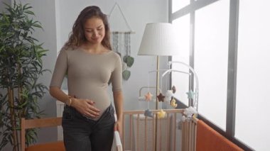 Bebek beşiği ve yumuşak oyuncaklarla çevrili, rahat bir bebek odasında karnını nazikçe kucaklayan hamile kadın, evde huzurlu annelik anları yakalıyor..