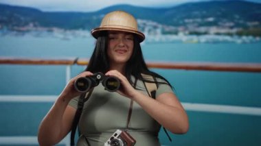 Uzun saçlı ve şapkalı genç bir kadın deniz kenarında dürbün tutuyor ve boynunda bir kamerayla rahat bir turist deneyimi sergiliyor..