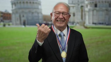 Takım elbiseli, altın madalyalı kıdemli bir adam başparmağıyla işeme kulesinin dışında gülümsüyor, parlak bir günde gururlu bir ifadeyle başarılı bir anı yakalıyor..