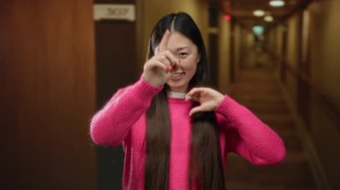 Pembe kazaklı kadın otel koridorunda kalp jesti yapıyor, uzun saçlarıyla gülümsüyor, ev ortamında kendine güveni ve cazibesini sergiliyor, dost canlısı atmosferi vurguluyor..