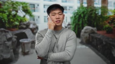 Genç adam, yeşil bitkiler ve modern binalarla dolu bir caddede yürürken akıllı bir telefonla konuşuyor, dinamik bir şehir manzarasında şehir yaşamını gözler önüne seriyor..