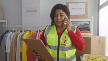 Yansıtıcı yelek giyen bir kadın yardım derneği bağış merkezinde elinde bir panoyla konuşuyor. Kıyafetler ve kutular organizasyon ve yardım çalışmalarına karıştığını gösteriyor..