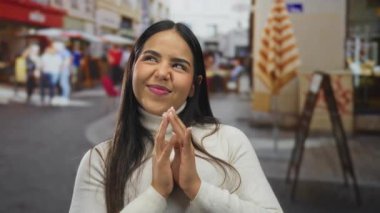 Kendine güveni tam bir kadın, neşeli bir caddede entrika çevirirken ellerini bir araya getiriyor. Genç Latin bir şahsiyet açık havada muzip, gülümseyen bir ifadeyle duruyor..