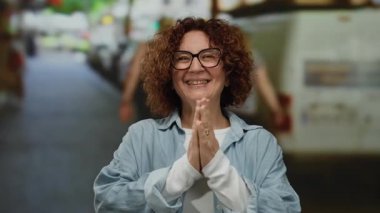 Kıvırcık saçlı ve gözlüklü, arka planı bulanık bir şehirde gülümseyen bir kadın.