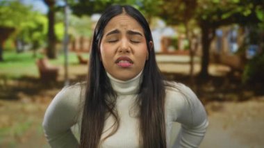 Kadının yüzü şaşkın görünüyor. Parkta genç Latin Latin kıyafetleri var. Açık havada yeşil giyinmiş, hafifçe eğiliyor..