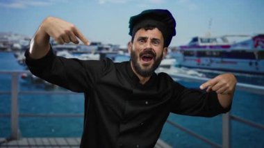 Siyah üniformalı bir şef gibi giyinmiş İspanyol sakallı adam arka planda gemiler ve teknelerle dolu bir limanın yakınlarında neşeli bir şekilde el kol hareketi yapıyor..