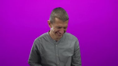 Canlı pembe arka planda gülümseyen bir adam, gözleri kapalı, neşeli bir ifade ve kapalı bir iç mekanda güçlü ışıklandırmayla günlük bir stil yakalıyor..