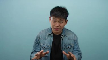 Kot ceket giyen genç Çinli adam, izole edilmiş mavi arka plana karşı açık bir şekilde jest yapıyor. Odaklanmış bir ifadeyle tartışma veya açıklama ima ediyor..