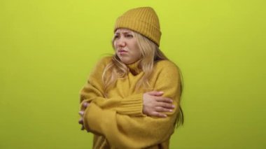 Sarı süveter ve bereyle yeşil arka planda titreyen kadın, kollarını sıkıca kavuşturarak soğuk rahatsızlığını dile getiriyor, kış modasını ve mevsimsel serinliği sergiliyor.