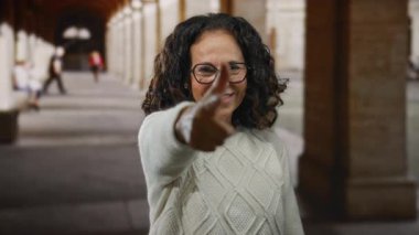 Kıvırcık saçlı bir kadın gündüz vakti üniversite kampüsünün önünde şakacı hareketlerle hareket ediyor.