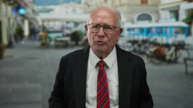 İş elbisesi giymiş kıdemli bir adam, hareketli bir açık hava caddesinde, bulanık binaları ve hareketli bir şehir geçmişi ile tutkuyla el kol geziyor..