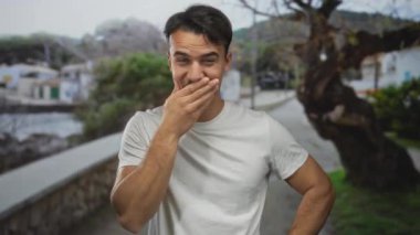 Genç İspanyol adam gülüyor ve deniz kenarındaki gezinti güvertesinde ağaçlar ve denizle birlikte rahat ve neşeli bir açık hava manzarasını canlandırıyor..