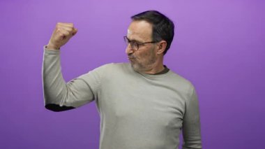 Latin kökenli yaşlı bir adam mor arka planda kollarını esnetiyor. Gözlük takıyor. Güç ve kendine güven gösteriyor..