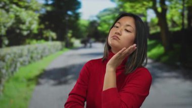 Kırmızı kazaklı genç bir kadın güneşli bir açık hava parkında başını uzatıyor yemyeşil ve arka planda bir patika var..