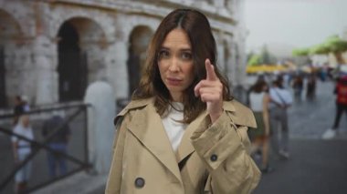 İtalya 'daki simgesel Roma kolezyumunun dışında bej rengi bir ceketle el kol hareketi yapan bir kadın, tarihi bir şehir ortamında turistlerin arasında merakını dile getiriyor..