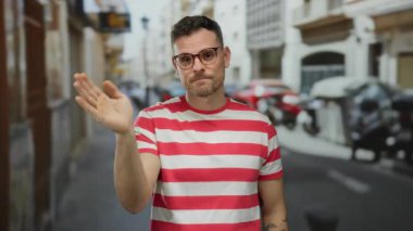 Çizgili tişörtlü genç adam güneşli gökyüzünün altında gözlüklü ve sıradan görünümlü bir şekilde şehir sokağında el sallıyor.