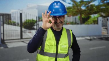 Miğferli ve yelekli bir adam gülümsüyor ve şehir dışındaki açık hava ortamında 