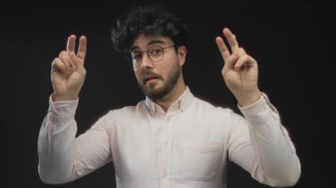 Sakallı ve gözlüklü genç bir adam siyah bir arka plana, bir stüdyo ortamında yüz ifadesine ve jestlere karşı tırnak işareti yapıyor..