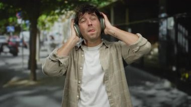 Güneşli bir sokakta kulaklıkla müzik dinleyen İspanyol bir adam, dışarıda keyif ve rahatlama ifade ediyor, şehir özgürlüğünün ve huzurunun özünü yakalıyor..