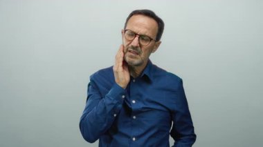 Beyaz bir arka plan karşısında diş ağrısı çeken İspanyol bir adam, çenesini acılı bir ifadeyle sıkıca tutuyor, yetişkin bir ortamda izole edilmiş diş rahatsızlığı gösteriyor..