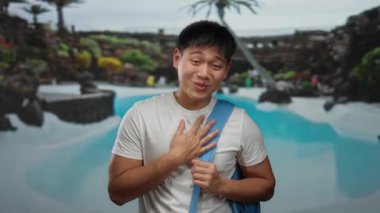 Sırt çantalı genç adam lüks havuzda sıcacık gülümsüyor. Bölgeyi çevreleyen tropikal bitkilerle dolu tesiste sakin bir atmosfer yaratıyor.