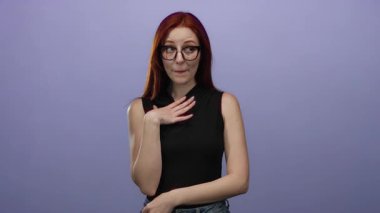 Kızıl saçlı ve gözlüklü bir kadın mor bir arka plana karşı duruyor, siyah bir bluz giyiyor, elleri göğsünde anlamlı bir jest yapıyor, güzelliğini ve tarzını ifade ediyor..