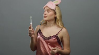 Gri arka planda diş fırçası tutan, pembe uyku maskesi takan ve diş fırçasını yakından inceleyen sarışın genç bir kadın.