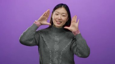 Elleriyle gülümseyen bir kadın canlı mor bir arkaplana karşı yüzünü çerçeveliyor rahat gri bir kazak giyiyor, sıcaklık ve kendine güven saçıyor..