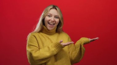 Sarışın bir kadın, izole edilmiş kırmızı bir arka planda mutlu bir şekilde el kol hareketi yapıyor sarı bir kazak giyiyor, sevinç ve heyecanı açık elleriyle ifade ediyor..