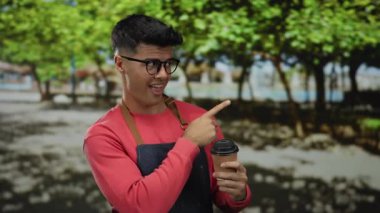 Parkta gülümseyen yakışıklı adam elinde kahve, kırmızı gömlek ve gözlüklerle açık havayı gösteren yeşil ağaçlarla çevrili parlak güneşli bir günü yansıtıyor..