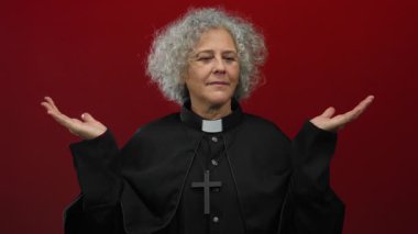 Gri saçlı, rahip kıyafeti giyen, kırmızı arka planda el ele tutuşan kadın sakin ve otoriter görünüyor.